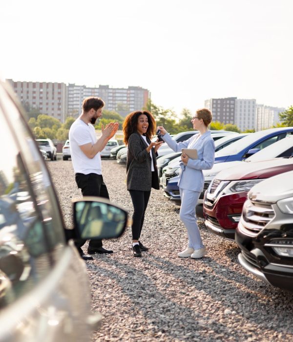 A car salesperson hands the keys to a new car owner as their partner claps in the background. They are at a car dealership.